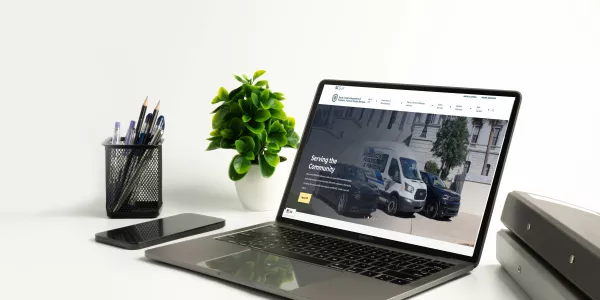 A clean white desk setup with an open laptop displaying the SCDPPPS homepage, a smartphone beside it, a small potted plant, a pen holder with pens, and stacked binders on the right against a white background.