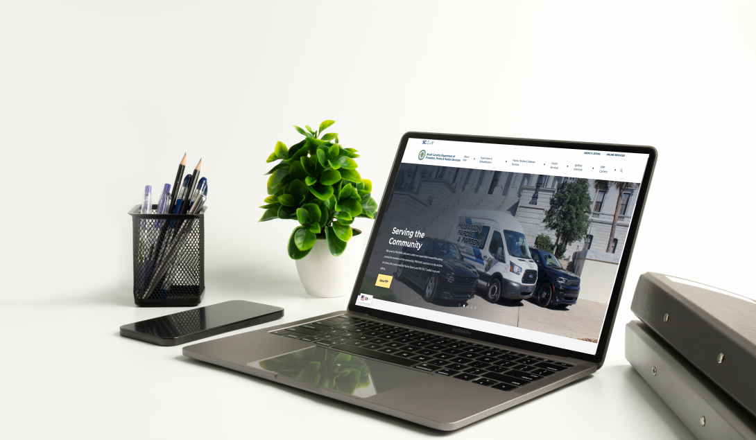 A clean white desk setup with an open laptop displaying the SCDPPPS homepage, a smartphone beside it, a small potted plant, a pen holder with pens, and stacked binders on the right against a white background.