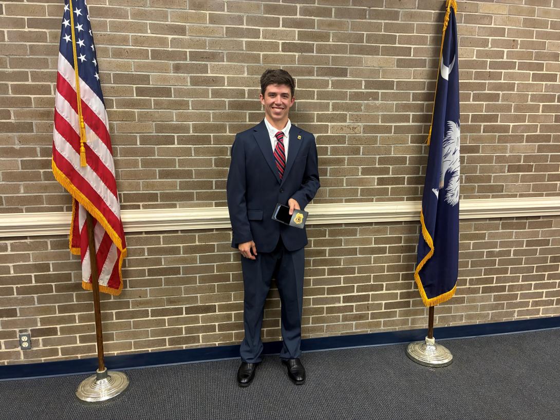 Man in a suit standing between the American flag and Palmetto flag..